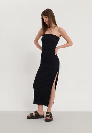 Robe maxi noire sans bretelles en tricot avec un motif texturé et une fente sur le côté, associée à des sandales noires épaisses avec des boucles.