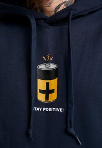 Sudadera azul marino con un gráfico de batería en negro y amarillo. El texto dice "¡MANTENTE POSITIVO!" debajo del diseño. Tejido suave, capucha ajustable.
