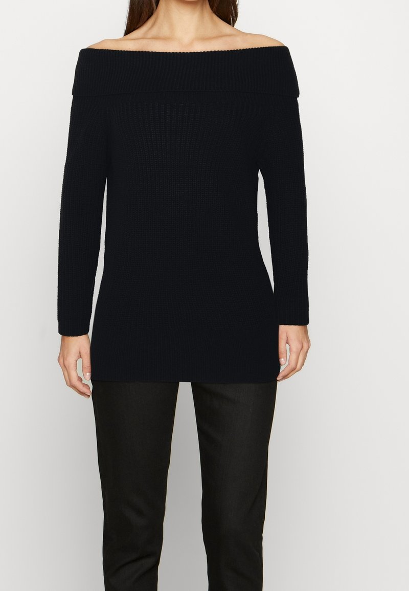Femme portant un pull côtelé noir à épaules dénudées avec des manches longues et un pantalon noir, debout devant un fond blanc uni.