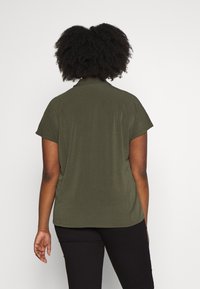 Vihreä oliivi lyhythihainen paita, jossa on kaulus, sileä rakenne, rento malli ja kerrostettu selkäosa, yhdistettynä mustiin housuihin.