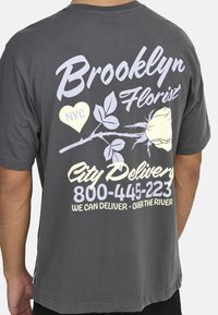 Grå bomulds t-shirt med et stort blomsterdesign, tekst i hvid og pastelfarver, samt leveringsinformation på bagsiden.