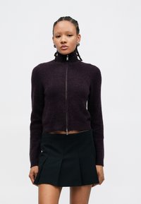 Vijoličen skrajšan pulover z visokim ovratnikom in sprednjim zadrgo, kombiniran s črno gubano krilom. Mehka tekstura, oprijeta zasnova.