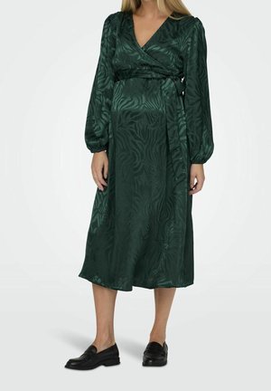 Woman wearing a long-sleeve, dark green, patterned wrap dress with a tied waist, paired with black loafers on a plain background.