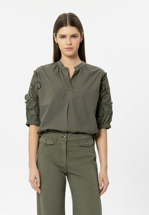 Junge Frau in olivgrüner Bluse mit floralen Ausschnitt-Ärmeln und passenden hoch taillierten Hosen, stehend vor einfachem weißem Hintergrund.