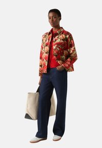 Jacke mit Blumenmuster in Rot, Grün und Creme, kombiniert mit einem roten Top, dunkelblauen Jeans mit weitem Bein und weißen Turnschuhen, dazu eine Leinwand-Shoppertasche.