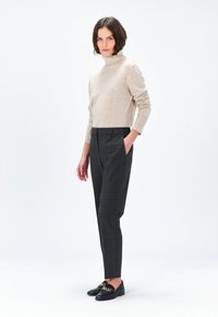 Maglione beige a collo alto realizzato in morbido materiale, abbinato a pantaloni sartoriali grigio scuro con motivo e mocassini neri con dettaglio in catena dorata.
