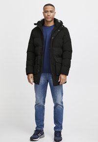Chaqueta de plumas negra con capucha, que cuenta con un cierre de cremallera y broches a presión, dos bolsillos delanteros, llevada sobre un sudadera azul, combinada con jeans de un azul claro.
