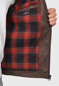 Veste en cuir marron avec une doublure en tartan rouge et noir, dotée d'une fermeture éclair et d'une étiquette de poche intérieure. Texture lisse sur le matériau extérieur.