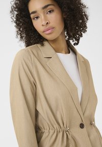 Hellbrauner Blazer mit einem einreihigen Knopfverschluss, taillierter Taille, geschlitzten Revers und langen Ärmeln; glatte Stofftextur mit dezentem Glanz.
