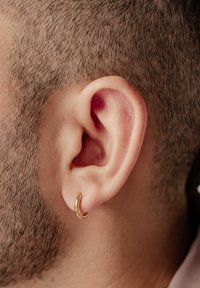 Orecchino a cerchio d'oro indossato in un orecchio forato, con una superficie liscia e un design circolare. Primo piano dell'orecchio che mostra capelli corti e pelle dettagliata.