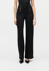 Γυναίκα που φοράει μαύρο παντελόνι με φαρδιά μπατζάκια και ψηλή μέση, με έξι χρυσά κουμπιά στο μπροστινό μέρος, συνδυασμένο με μαύρες μυτερές γόβες.