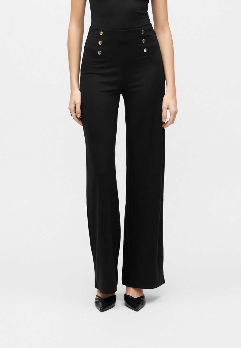 Γυναίκα που φοράει μαύρο παντελόνι με φαρδιά μπατζάκια και ψηλή μέση, με έξι χρυσά κουμπιά στο μπροστινό μέρος, συνδυασμένο με μαύρες μυτερές γόβες.