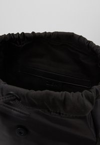 Bolsa de drawstring negra con una textura suave, parte superior recogida y forro liso. El interior presenta un simple detalle de etiqueta negra.