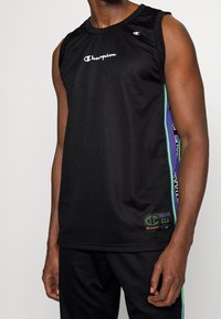 Černý sportovní top Champion bez rukávů z prodyšné síťoviny s bílým logem na hrudi a zelenými a fialovými pruhy po stranách, který nosí osoba s tmavou pletí.