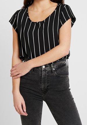 Young woman in black short-sleeve top with white vertical stripes and high-waisted faded black jeans, standing with arms crossed.