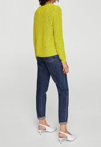 Helle gelbe Strickjacke mit Strukturmuster, kombiniert mit hochgekrempelten dunkelblauen Jeans und weißen Absatzschuhen.