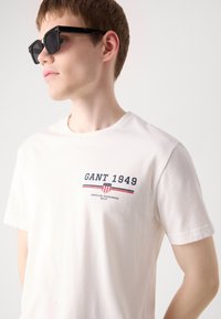 Hvid bomulds t-shirt med "GANT 1949" logo i marineblå og rød. Rund hals, korte ærmer, blød tekstur, afslappet pasform.