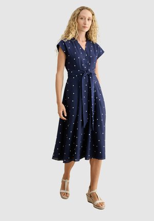 Frau mit lockigem blondem Haar, die ein marineblaues Midikleid mit weißen Punkten und gebundener Taille sowie beigefarbene Keilsandalen trägt, steht vor weißem Hintergrund.