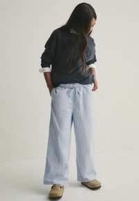 Helle Baumwollhose mit weitem Bein in blau-weiß gestreift, elastischem Bund, Seitentaschen, kombiniert mit einem dunklen Sweatshirt und beigen Schuhen.