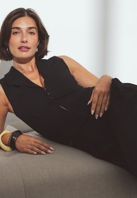 Mustmusthüpseline kombinesoon, millel on kaelus ja eeslukuga, suurte ringikujuliste käevõrudega, kerge halli tausta vastas.