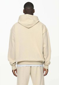 Beige oversized hettegenser med snøring. Mykt, teksturert stoff. Ribbestrikkede mansjetter og kant. Kombinert med matchende beige joggebukser.