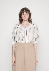 Blusa branca com riscas verticais em azul, castanho e laranja, com mangas bufantes e decote redondo. Usada com calças bege de cintura alta.