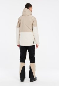 Giacca impermeabile beige e bianca con cappuccio, indossata sopra pantaloni neri con accenti beige. Presenta dettagli con zip e tessuto testurizzato.