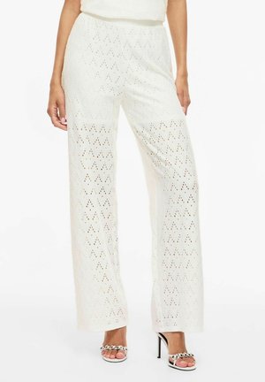 Femme portant un pantalon blanc en maille à jambes larges avec un motif en losange, associé à des sandales blanches à talons hauts avec des ornements argentés.