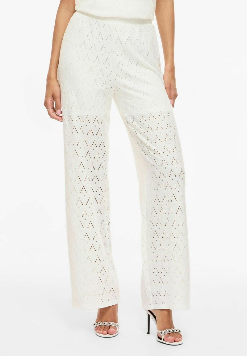 Femme portant un pantalon blanc en maille à jambes larges avec un motif en losange, associé à des sandales blanches à talons hauts avec des ornements argentés.
