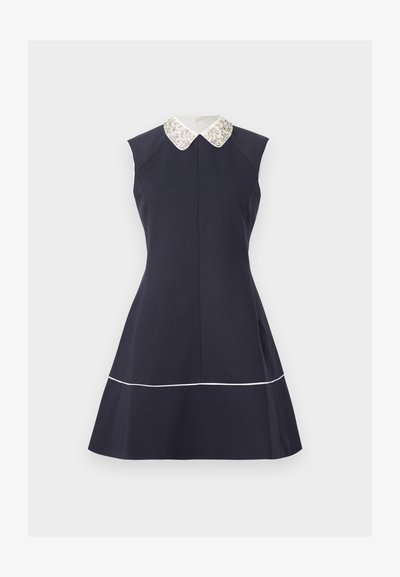 Φόρεμα χωρίς μανίκια σε ναυτικό μπλε χρώμα με φαρδιά φούστα, λευκό κολάρο τύπου Πίτερ Παν με διακόσμηση και λεπτό λευκό τελείωμα κοντά στη ραφή, σε ανοιχτό φόντο.