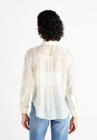 Camisa blanca ligera con una textura transparente, un sutil patrón de lunares y mangas largas. Cuenta con un cuello abotonado y un detalle en la pieza trasera.