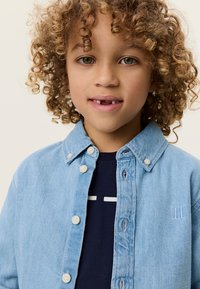 Ανοιχτό μπλε τζιν πουκάμισο με σχέδιο με κουμπιά, δύο μπροστινές τσέπες και ένα ναυτικό μπλουζάκι από κάτω με λευκό γραφικό μοτίβο.