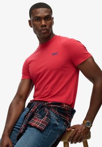 Camiseta de algodón roja con cuello redondo y mangas cortas, que presenta un pequeño logo azul. Combinada con unos vaqueros azules y una camisa de cuadros atada a la cintura.
