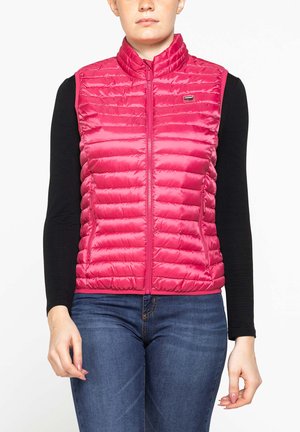 Donna che indossa un gilet trapuntato smanicato di colore rosa acceso sopra una maglia nera a maniche lunghe e jeans blu, in piedi davanti a uno sfondo bianco.