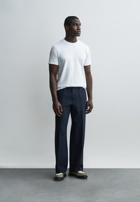 Uomo con camicia bianca a maniche corte, pantaloni blu navy e mocassini bicolore in piedi davanti a una parete grigia chiara liscia.