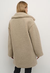 Beige, pörröinen lampaanvillatakki, jossa on suuri kaulus ja leveät hihat, rento istuvuus ja pehmeä rakenne, joka ulottuu reiden keskikohtaan.