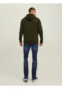 Felpa verde oliva con cappuccio regolabile, maniche lunghe e polsini a costine, abbinata a jeans blu scuro e sneaker navy.
