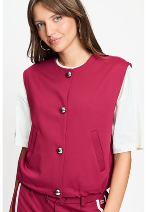 Jonge vrouw die een mouwloos magentakleurig vest met zilveren knopen draagt over een wit T-shirt met korte mouwen, poserend tegen een effen achtergrond.