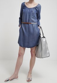 Nainen, jolla on päällä sininen farkkukontrasti mekko ruskealla vyöllä, hopeiset liukuva sandaalit ja hän kantaa isoa teksturoitua hopeista käsilaukkua.