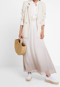 Lange, vloeiende beige rok van lichtgewicht stof, gecombineerd met een witte blouse en een lichte blazer. Accessoires omvatten een geweven rieten tas en witte sneakers.
