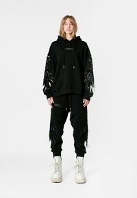 Conjunto de sudadera negra y joggers con embellishments similares a plumas en las mangas y los laterales del pantalón, con un acabado texturizado y botas blancas con cordones.