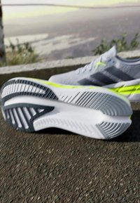 Zapatillas de correr gris claro con acentos en amarillo neón sobre un pavimento, una zapatilla mostrando la suela texturizada, entorno al aire libre con fondo difuminado.