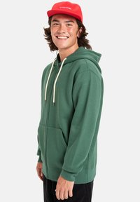 Groene katoenen hoodie met een ritssluiting aan de voorkant, koordjes en een kangoeroezak. Draagt een rode baseballcap met een logo.