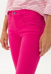 Pantalones ajustados de color rosa brillante con bolsillos frontales y detalles de costura sutiles, combinados con una blusa a rayas. Textura suave, tela ligera.