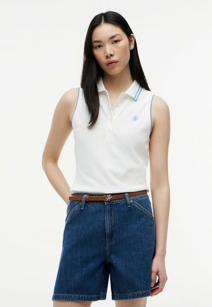 Ung kvinde med langt mørkt hår iført en hvid ærmeløs poloshirt og blå denimshorts med et brunt bælte foran en ensfarvet baggrund.