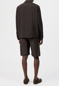 Mann iført mørk langermet skjorte, matchende knelang shorts og mørke casual sko, stående med ryggen mot på ensfarget hvit bakgrunn.