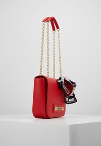 Punainen nahkainen käsilaukku, jossa on neliömäinen muoto, kultaiset yksityiskohdat ja kaksinkertaiset ketjuhihnat. Mallilla on yksi hihna sidottuna kuvioitu huivi koristeena.