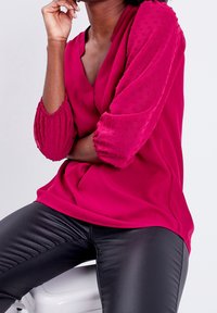 Blusa rosa vibrante con mangas abullonadas texturizadas, escote en V profundo y tela suave que cae sobre un par de pantalones negros ajustados.