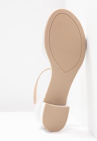 Semelle de sandale blanche avec semelle en caoutchouc beige et petit talon bloc, taille indiquée 4½, positionnée verticalement contre un fond blanc et un coin.