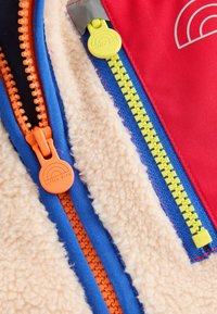 Kolorowa kurtka z czerwoną warstwą zewnętrzną i kremowym futerkiem wewnątrz, z kontrastującymi zamkami w kolorach pomarańczowym, żółtym i niebieskim oraz logo "Little Bird".